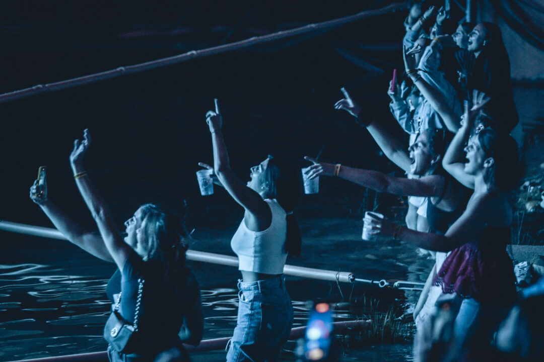 Grupo de asistentes bailando y levantando los brazos junto al agua en un concierto nocturno del Festival Pirineos Sur.